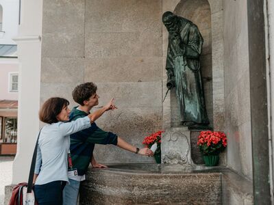 Stadtführung Altötting's Brauchtümer, Foto Tourismusverband Inn-Salzach Eine Stadtführerin steht mit einem Gast vor dem Bruder Konrad Brunnen, der sich gerade eine Flasche Wasser abfüllt.