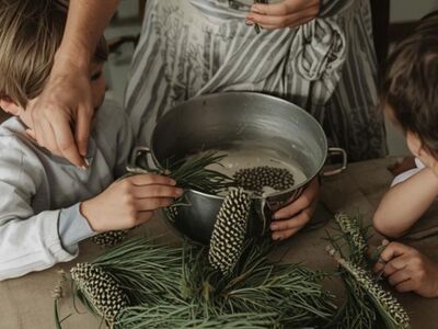 Eine Frau und zwei Kinder stehen vor einem Topf mit Nadeln und Tannenzapfen.