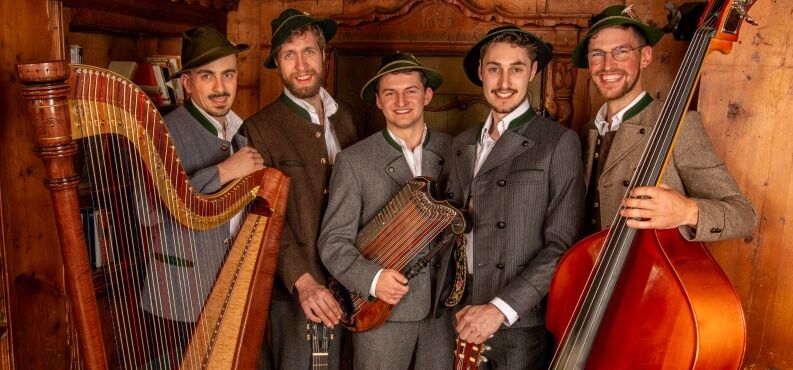 Fünf Musiker in Tracht stehen mit ihren Instrumenten vor einer Holzwand.