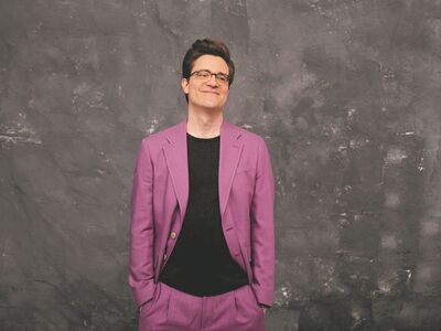 Ein Mann in lilafarbenem Anzug vor dunklem Hintergrund.