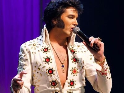 Elvis, Jimmy Holmes, forum Altoetting, Europolis, Bild Veranstalter Ein Mann in einem weißen Glitzeranzug hält ein Mikrofon in der Hand und singt.