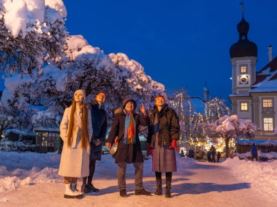 Führung Herzerwärmendes Altötting, Foto Dirschl Vier Frauen stehen auf dem verschneiten Kapellplatz bei Abenddämmerung.