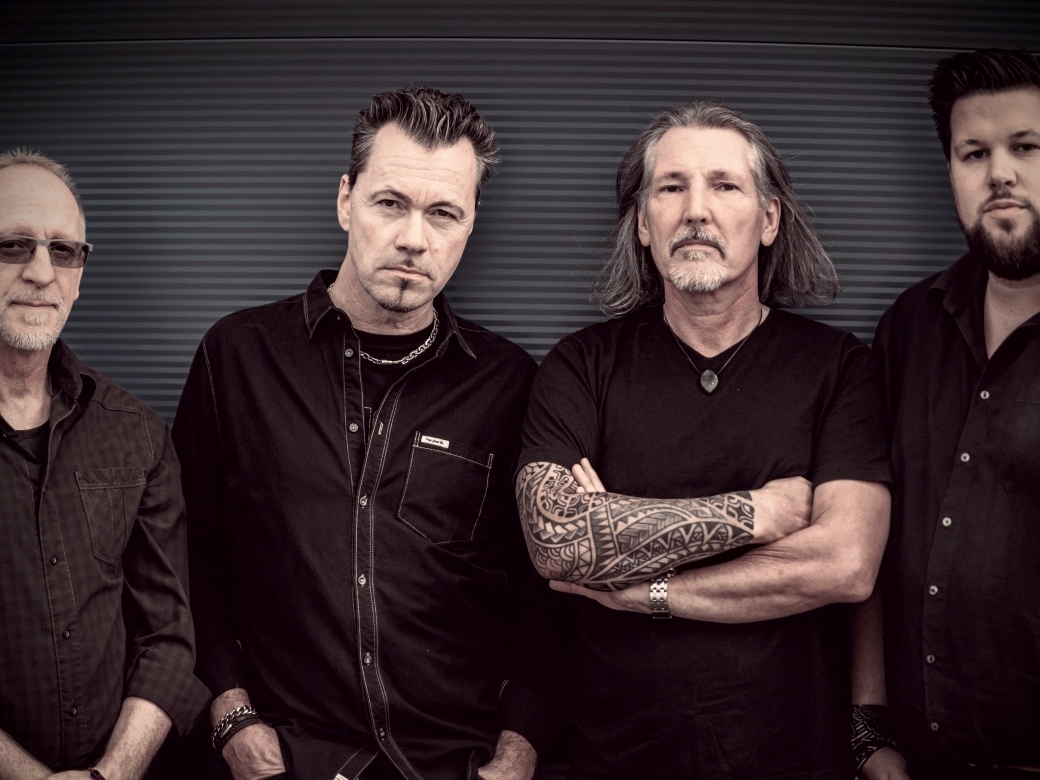 Vier Männer in schwarzen Shirts stehen vor einer grauen Wand.