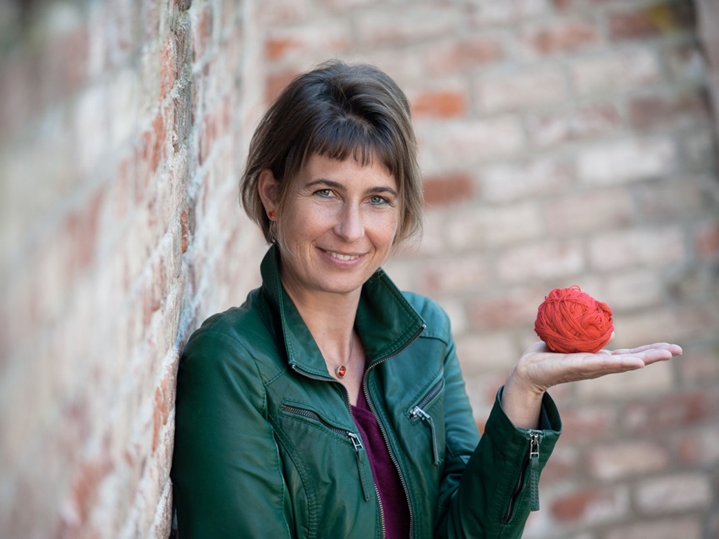 Eine Frau in grüner Jacke steht vor einer Steinmauer.
