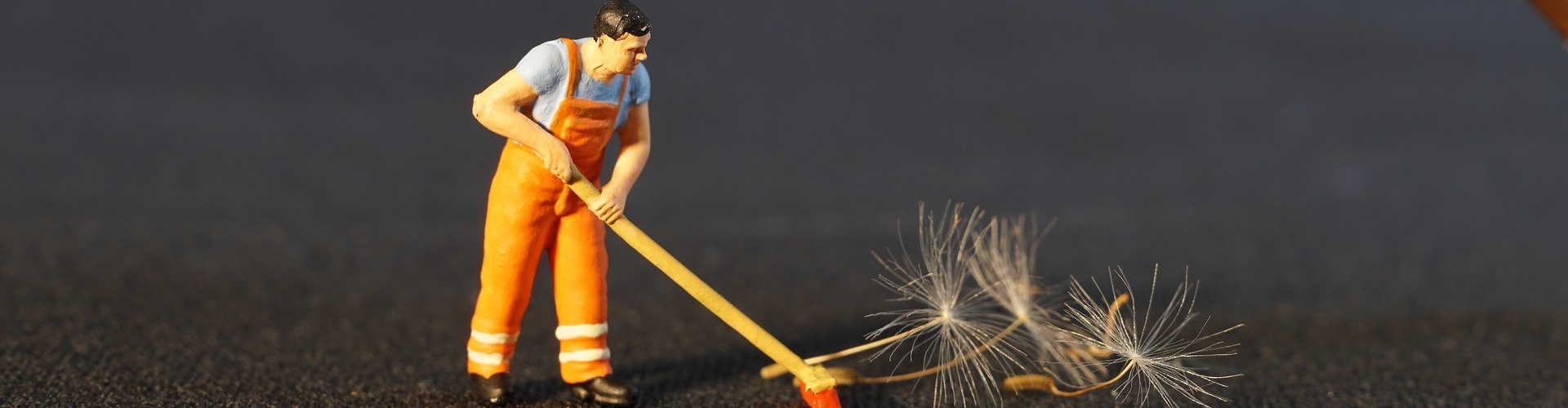 Ein Figur gekleidet als Bauhofmitarbeiter fegt die Samen einer reifen Löwenzahnpflanze weg, die im oberen Bildrand zu sehen ist. 