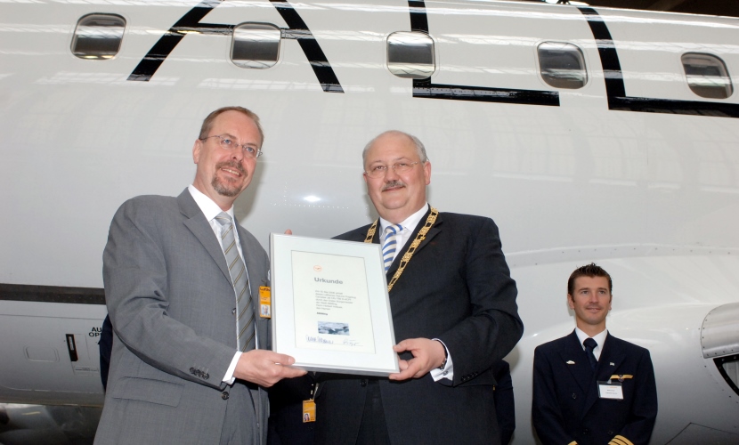Bürgermeister Hofauer und der Lufthansa Chef halten die Urkunde vor dem Flugzeug.
