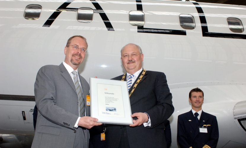 Bürgermeister Hofauer und der Lufthansa Chef halten die Urkunde vor dem Flugzeug.