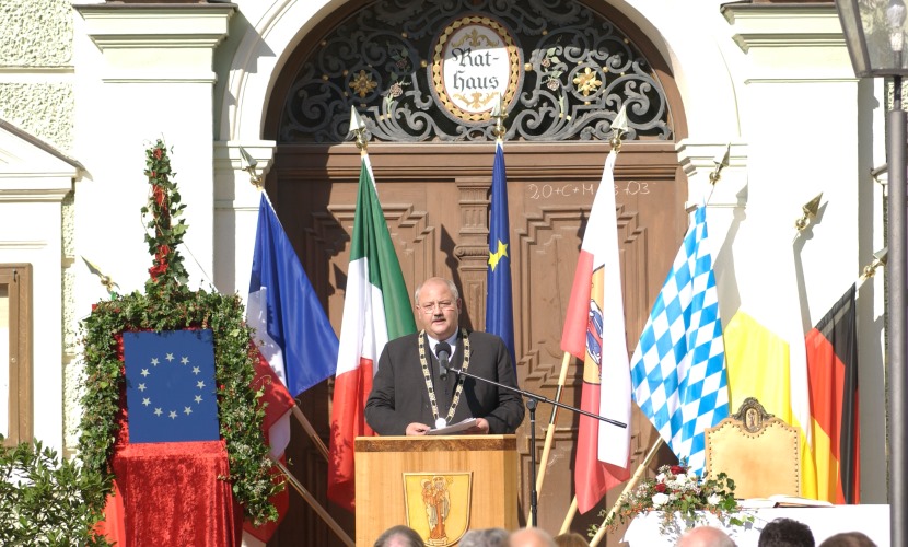 Bürgermeister Hofauer spricht am Rednerpult mit Fahnen im Hintergrund vor dem Rathaus. 