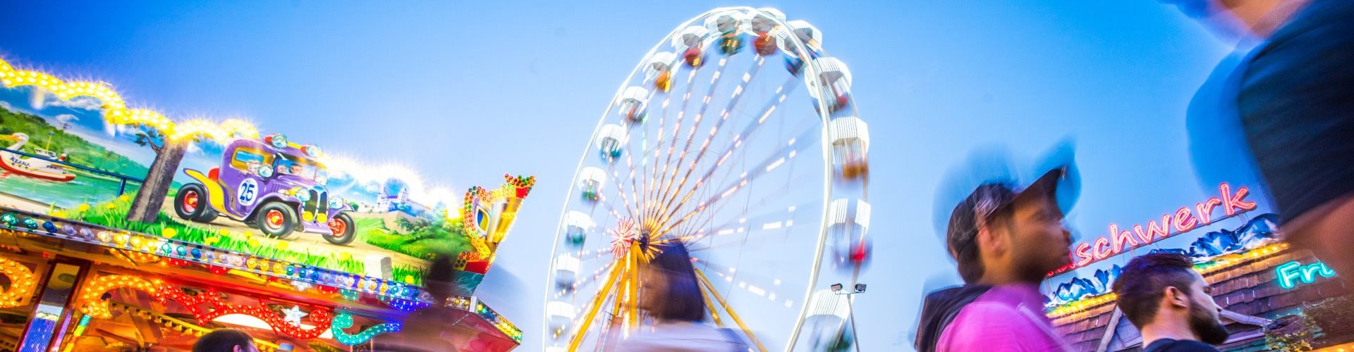 Besucher vor den Fahrgeschäften und dem Riesenrad auf der Hofdult Altötting in Abenddämmerung.