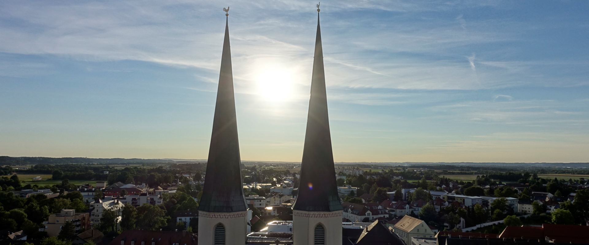 Luftaufnahme der Türme der Stiftspfarrkirche, zwischen denen die Sonne durchscheint.