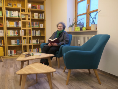 Klassiker Lesung in der Stadtbücherei Altötting, Foto Bücherei Eine Dame sitz mit einem Buch in der Hand auf einem Sessel vor einem Bücherregal.