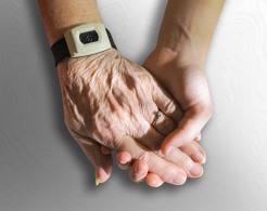 Eine junge Hand hält die Hand eines alten Menschen mit einem Hausnotrufknopf am Handgelenk.