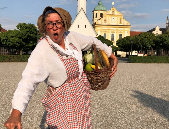 Eine alte Marktfrau steht mit ihrem Obstkorb auf dem Kapellplatz