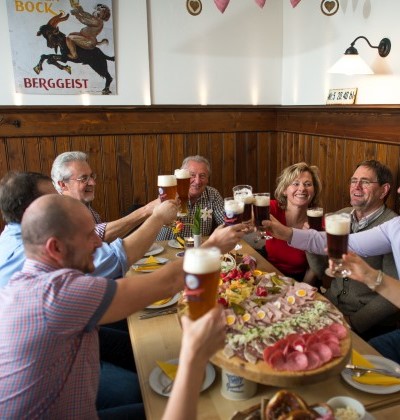 Menschen sitzen im Wirtshaus bei Brotzeit gemütlich zusammen und stoßen mit einem Bier an. 