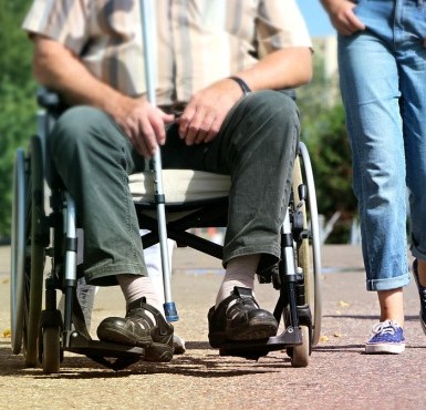 Ein gehbehinderter Mensch sitzt im Rollstuhl und wir von einer Person begleitet. 