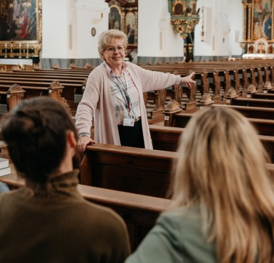 Zwei Frauen sitzen in der Kirchenbank und die Stadtführerin erzählt etwas.