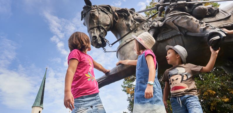 Hier sehen Sie 3 Kinder vor der Tilly Statue am Altöttinger Kapellplatz