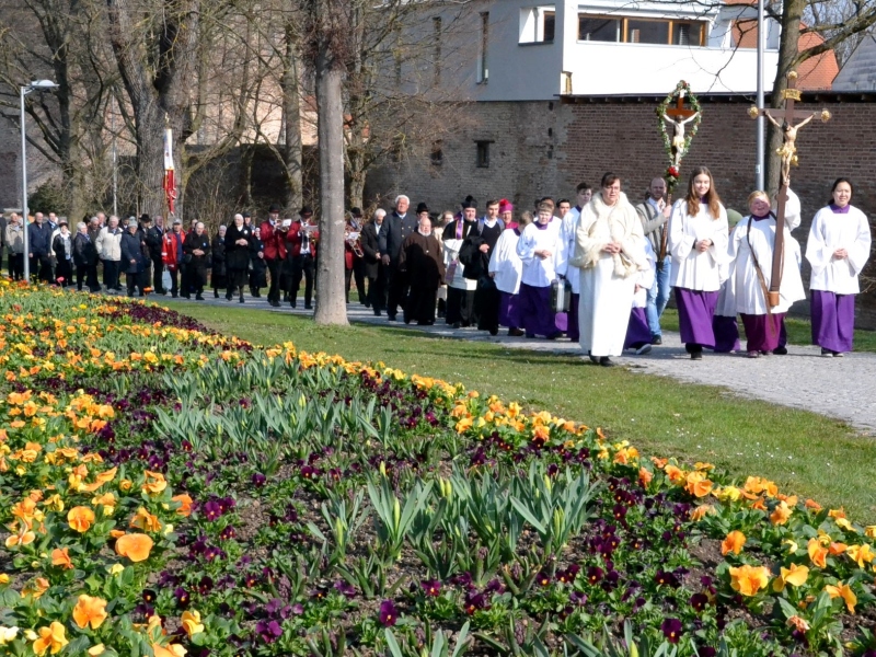 Die Geistlichkeit und die Pilger ziehen in Straubing in die Basilika ein mit Blumenbeet im Vordergrund. 