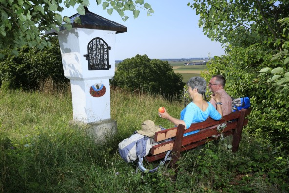 Ein Paar, das auf dem Rupertweg Richtung Altötting Rast auf einem Bänkchen vor einer Kreuzweg Station macht.