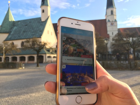 Ein Handy mit der offenen App mySpots am Altöttinger Kapellplatz mit Blick auf die Gnadenkapelle und die Stiftspfarrkirche.