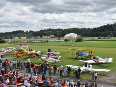 Kleinflugzeuge und Propellermaschinen stehen auf der Wiese mit Besuchern.