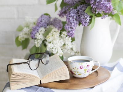 Auf einem Tisch liegt ein offenes Buch, eine Brille, ein Kaffee und eine Blumenvase mit Flieder.