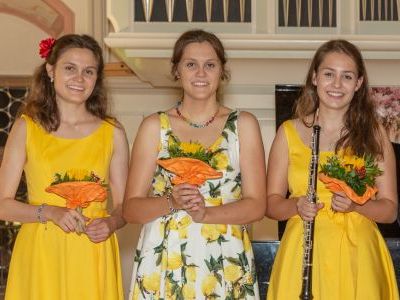 Drei Damen in gelben Kleidern halten jeweils einen Blumenstrauß.